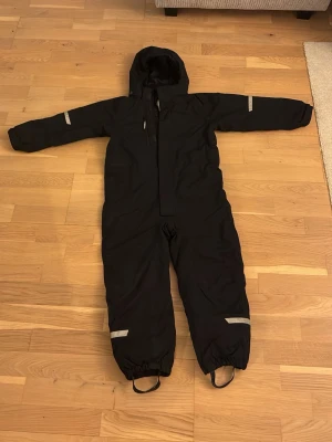 Svart täckoverall från Polarn O. Pyret - Svart vinteeoverall från Polarn O. Pyret med huva, dragkedja och reflexdetaljer på armar och ben. Tillverkad i slitstarkt, vind- och vattentätt material med återvunnen vaddering och foder. Perfekt för kalla dagar och vinteraktiviteter.
