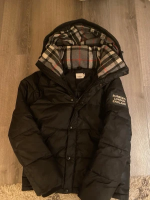 Svart pufferjacka från Burberry - Säljer en svart pufferjacka från Burberry med stor huva och klassiskt rutigt foder i grått, vitt, svart och rött. Jackan har en tydlig Burberry London England-patch på ärmen, dragkedja och knappar framtill. Perfekt för kalla dagar och riktigt snygg streetstil.