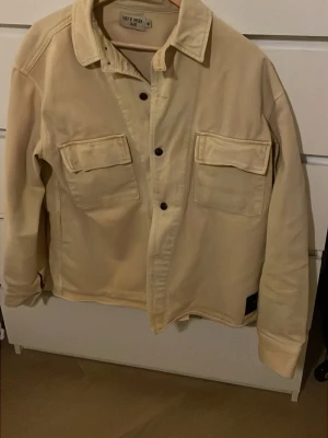 Beige overshirt från Tiger of Sweden - Säljer en stilren beige overshirt från Tiger of Sweden Jeans. Jackan har två stora bröstfickor med lock, klassisk krage och knäppning framtill. Perfekt för lager-på-lager och enkel att matcha med olika outfits.