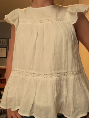 Vit blus med volang och spetsdetaljer - Söt vit blus i luftig bomull med volangärmar och fina spetsdetaljer vid bröstet och midjan. Blusen har en lös passform och är perfekt för varma dagar. Den har en romantisk och somrig känsla med sina mjuka rynk och tunna tyg.