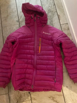 Peak Performance dunjacka M - Säljer en snygg rosa/lila dunjacka från Peak Performance med huva. Modellen heter Heli down jacket med quiltad design, hel dragkedja framtill, förstärkt på axlar, bröstficka och snörning i nederkant. Perfekt för kyliga dagar, skidåkning och funkar även bra framåt våren och riktigt skön att ha på sig. Nästan ny använd fåtal gånger.