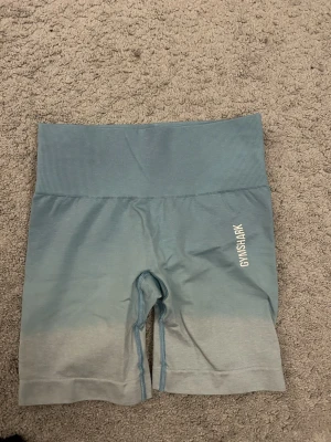 Blå shorts från Gymshark - Snygga blå shorts från Gymshark i storlek XS. De har hög midja och en ombre-effekt som går från ljusblått till grått. Perfekta för träning och har en tight passform som sitter på plats under hela passet.