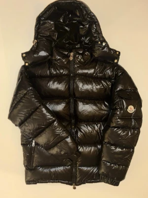Svart Moncler pufferjacka med huva - Säljer en svart, glansig pufferjacka från Moncler med huva och dragkedja framtill. Jackan har en ficka på ärmen med Moncler-logga och två sidofickor med dragkedja. Insidan har ett coolt serietryck. Perfekt för kalla dagar och riktigt snygg streetstil. Jätte fina skick hör av er vid funderingar pris är inte hugget i sten, storlek m passar kanske L