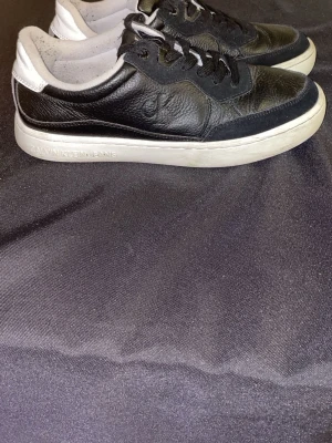 Svarta sneakers från Calvin Klein Jeans - Säljer ett par svarta sneakers från Calvin Klein Jeans med vit sula och CK-logga på sidan och plösen. Skorna har snörning, rund tå och är tillverkade i skinn och syntet. Perfekta för dig som gillar stilrena och klassiska sneakers.