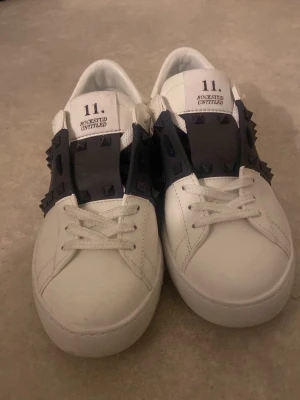 Valentino Rockstud Untitled sneakers - Säljer ett par vita Valentino Rockstud Untitled sneakers i skinn med svarta detaljer och ikoniska nitar över vristen. Skorna har rund tå, vita snören och en chunky sula. Perfekta för dig som vill ha en edgy och lyxig touch till din outfit. Aldrig använda