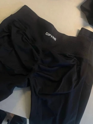 Svarta tights från DFYNE - Säljer ett par svarta tights från DFYNE med hög midja och ribbad linning. Materialet är stretchigt och passar perfekt för träning eller chill. Snygg scrunch-detalj bak för extra form. Supersköna och sitter på plats hela passet.
