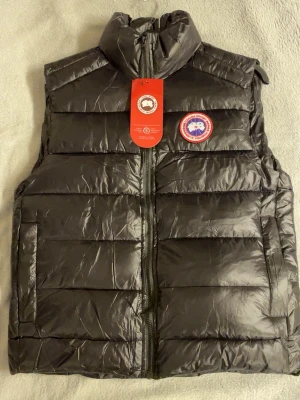 Svart dunväst från Canada Goose - Säljer en svart dunväst från Canada Goose med hög krage och dragkedja framtill. Västen har det klassiska runda Canada Goose-märket på bröstet och är quiltad med breda paneler. Perfekt för lager-på-lager och riktigt snygg till höst och vinter.