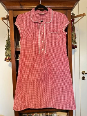 Rödvit ginghamklänning - Röd och vit retro gingham klänning med fina lace spets detaljer och hjärtformade knappar. Finns en liten ficka på ena brösten och två fickor i sidan. Den är som ny och har inga slitage och är helt oanvänd av mig. Säljer då den tyvärr är för stor:( Hittar faktiskt ingen storlek men personen jag köpte den av hade den uppe som en XS. Priset är förhandlingsbart och bara skriv om ni har några frågor!