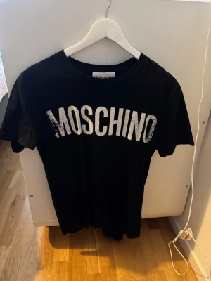 Svart Moschino t-shirt med logga - Säljer en svart t-shirt från Moschino Couture med stor vit logga framtill. Klassisk rund halsringning och korta ärmar. Tillverkad i mjuk bomull för skön känsla och enkel stil. Perfekt för dig som gillar statement-plagg och italiensk design.