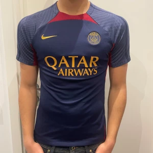 PSG matchtröja Nike marinblå/gul - Säljer en Paris Saint-Germain matchtröja från Nike i marinblått med gula detaljer och logga. Tröjan har röda inslag vid axlarna och mönstrade ärmar. Materialet är syntet och perfekt för fotbollsträning eller att bära som supporter. Inget tryck har lossnat.