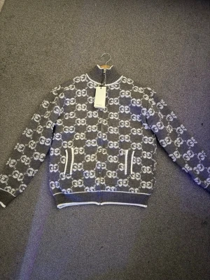 GG cardigan - Säljer en grå och vit kofta med ikoniskt GG-mönster över hela plagget. Tröjan har ribbad hög krage, dragkedja framtill och två fickor med vita detaljer. Perfekt för dig som vill sticka ut med en lyxig och trendig look. Fits Size S!!!!