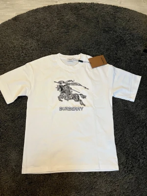 Vit Burberry t-shirt med tryck - Vit t-shirt från Burberry med ett stort svart tryck av en riddare på häst och texten 'BURBERRY' framtill. Klassisk passform med rund hals och korta ärmar. Perfekt för dig som gillar stilrena designer med en lyxig touch. Storlek L och aldrig använd, hör av dig vid funderingar!