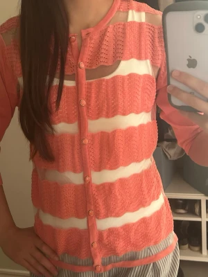 Korallfärgad kofta - Korall/orangerosa kofta med genomskinliga detaljer på framsidan. De genomskinliga delarna är tyg med glittrig tråd. Man kan ha olika toppar under koftan som charmigt syns igenom. Mycket somrig kofta som kan kläs upp och ned!  Storlek S/M   Märket Mudflower