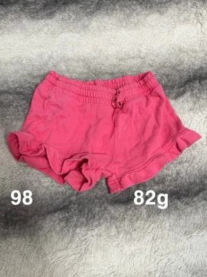 Rosa volangshorts med snörning - Säljer ett par söta rosa shorts med volangkant och snörning i midjan. De har elastisk midja för skön passform, perfekta för varma dagar.🌸 Fint använt skick. Storlek 98 