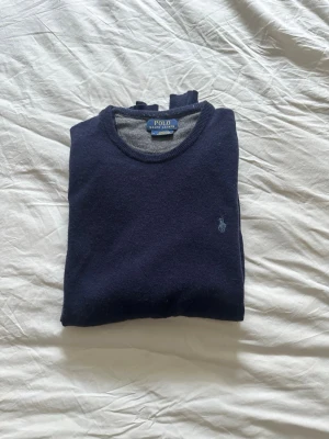 Mörkblå mjuk stickad tröja från Polo Ralph Lauren - Säljer en klassisk mörkblå tröja från Polo Ralph Lauren i mjuk Merino. Tröjan har rund halsringning, diskret logga broderad på bröstet och långa ärmar. Perfekt för dig som gillar stilrena och tidlösa plagg. Tröjan är i gott skick 