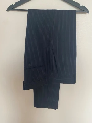 Mörkblå randiga kostymbyxor - Snygga mörkblå kostymbyxor med tunna vita ränder från Riley .Perfekt för en clean och stilren look. Ser ut som nya!!