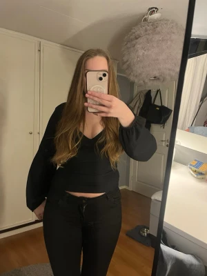 Svart blus med v-ringning och knyt - Säljer en svart blus med v-ringning och långa puffärmar. Toppen har croppad passform och en snygg öppen rygg med knytband upptill. Materialet är lätt strukturerat och känns mjukt mot huden. Perfekt för dig som gillar stilrena och trendiga plagg.