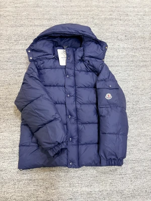 Mörkblå Moncler jacka  - Säljer en mörkblå dunjacka från Moncler med stor huva och klassisk logga på ärmen. Jackan har quiltad design, tryckknappar och dragkedja. Insidan har en cool serietidningsetikett och flera detaljer. Perfekt för dig som vill ha en varm och snygg jacka i vinter.