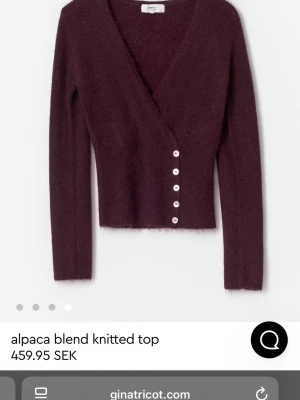 Vinröd omlottkofta från Gina Tricot - Snygg vinröd omlottkofta från Gina Tricot i mjuk alpackablandning. Koftan har v-ringning, långa ärmar och dekorativa knappar framtill. Perfekt för dig som gillar en stilren och mysig look med lite extra detaljer.