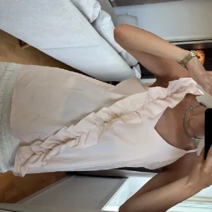 Ljusrosa linnetopp med volang - Säljer en ljusrosa linnetopp med bred volangdetalj som går diagonalt över framsidan. Toppen är ärmlös och har en v-ringad halsringning. Materialet känns lätt och luftigt, perfekt för varma dagar. Snygg och feminin design som ger en romantisk vibe. Liten fläck de bild två 