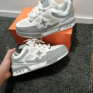 Louis Vuitton sneakers i vitt och grått - Snygga Louis Vuitton sneakers i vitt och grått med coola LV-detaljer och broderade loggor. Skorna har meshpaneler, skinnpartier och robust platt sula. Ikoniska monogramdetaljer på sidan och hälen, samt snörning framtill. Perfekta för dig som vill sticka ut med lyxig streetstyle. De är ett till ett och med perfekt kvalite.