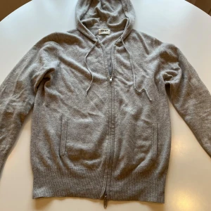 Grå kashmirhoodie från Soft Goat - Mjuk och stilren ljusgrå hoodie i 100% kashmir från Soft Goat. Tröjan har dragkedja framtill, huva med snörning och ribbade muddar. Perfekt för dig som vill ha en clean och lyxig look med maximal komfort. En liten defekt men man ser ej vid andvändning! Hör av dig vid minsta fundering ! MVH Leo