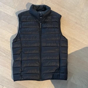 Svart dunväst från Uniqlo XS - Svart, quiltad dunväst från Uniqlo i modellen Ultra Light Down. Västen har hög krage, dragkedja framtill och är superlätt. Perfekt för lager-på-lager och enkel att packa ner i väskan. Klassisk och stilren design utan mönster.