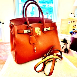 Brun handväska med gulddetaljer - Snygg brun handväska i skinn med klassisk fyrkantig form och guldfärgade metalldetaljer. Väskan har två handtag, avtagbar axelrem och lås framtill. Insidan är rymlig med en innerficka. Perfekt för dig som gillar stilrena accessoarer med lyxig känsla.