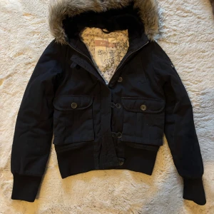 Svart pufferjacka med päls från Sublevel - Svart pufferjacka från Sublevel med fluffig päls på huvan och quiltat, ljusrosa foder. Jackan har stora fickor med knappar, dragkedja och extra knäppning framtill. Ribbad mudd vid ärmslut och nederkant. Perfekt för kalla dagar och har en cool, urban vibe.