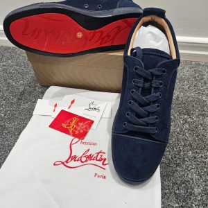Mörkblå sneakers från Christian Louboutin - Snygga mörkblå sneakers från Christian Louboutin i mocka med klassisk röd sula. Skorna har rund tå, snörning och diskreta detaljer i samma färg. Insidan är ljusbeige och sulan är platt. Perfekta för dig som vill ha något exklusivt och stilrent.