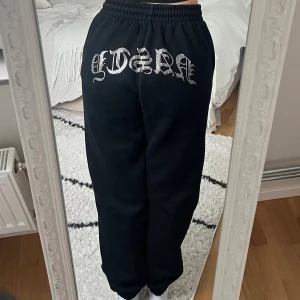 Svarta mjukisbyxor med strassdetalj 🖤 - Svarta Hidden Cult mjukisbyxor med elastisk midja och snörning framtill. Storlek S i fint skick. På baksidan finns en stor strassdekor med texten 'NASTY' i gotisk stil. Byxorna har en loose passform och är tillverkade i mjukt bomullsmaterial, perfekta för chill dagar. Har även tillhörande hoodie i en annan annons 🖤