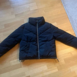 Svart pufferjacka med dragkedja - Svart pufferjacka med hög krage och snygga quiltade detaljer. Jackan har två stora fickor framtill, hel dragkedja och snörning i nederkant för att justera passformen. Perfekt för kalla dagar och har en clean, modern look. Står storlek XS, men passa också S!