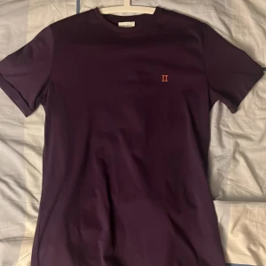 Lila t-shirt från Les Deux - Snygg lila t-shirt från Les Deux med korta ärmar och rund hals. På bröstet finns en liten broderad detalj med romerska siffran II i orange. T-shirten är gjord i mjuk bomull och har en clean, stilren look som funkar till många outfits.