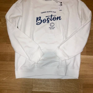 Vit Tennis World Cup Boston sweatshirt från 157 Laget - Vit sweatshirt från 157 med blått tryck på bröstet där det står 'Boston' och 'Tennis World Cup'. Tröjan har rund hals, långa ärmar och ribbade muddar. Perfekt för dig som gillar sportig stil och vill ha något bekvämt och snyggt.