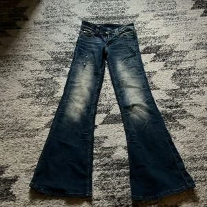 Bootcut jeans från Cheap Monday - Säljer ett par blå bootcut jeans från Cheap Monday med slitna detaljer och ljusa partier framtill. Klassisk femficksmodell med låg midja. Snyggt Cheap Monday-märke bak i svartvitt och orange. Säljer pågrund av att de är försmå och är köpta från Plick. 