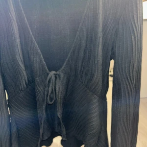 Svart ribbad omlottopp med knyt - Säljer en svart omlottopp med ribbad struktur och långa ärmar. Toppen har v-ringning och knyt framtill som ger en snygg siluett. Materialet är mjukt och stretchigt, perfekt för att skapa en cool look. Passar dig som gillar stilrena och trendiga plagg.