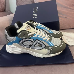 Sneakers med blå och grå detaljer - Säljer ett par snygga sneakers med chunky vit och svart sula, grå mesh, blå och vita paneler samt stora CD-loggor på sidan. Skorna har snörning och en modern, sportig vibe med coola kontraster och detaljer. Perfekt för dig som gillar exklusiva märken och streetstyle.