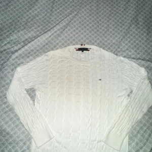 Vit kabelstickad tröja Tommy Hilfiger - Kabelstickad vit tröja från Tommy Hilfiger med rund halsringning och diskret logga på bröstet. Tröjan har långa ärmar och ribbade muddar. Perfekt för dig som gillar klassisk och stilren look.