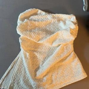 Vit axelbandslös topp - Säljer en vit axelbandslös topp med snygg struktur i tyget. Den har en omlottliknande överdel och är tillverkad i ett lätt, mönstrat material som ger en cool och modern vibe. Perfekt för dig som gillar stilrena och trendiga plagg.