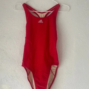 Röd baddräkt från Adidas, storlek M - Sportig röd baddräkt från Adidas med klassisk logga framtill och snyggt brottarrygg med öppen detalj. Tillverkad i stretchigt material med 78% nylon och 22% elastan, fodrad med 100% polyester. Perfekt för simträning eller strandhäng.