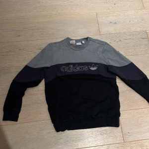 Grå och svart Adidas sweatshirt - Säljer en snygg Adidas sweatshirt i grått, svart och mörkblått med broderad logga framtill. Tröjan har rund hals, långa ärmar och är tillverkad i mjuk fleece på insidan. Perfekt för dig som gillar sportig och avslappnad stil.