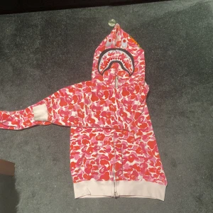 BAPE Shark Hoodie rosa camo - Säljer en BAPE Shark Hoodie i rosa, röd och vit camo bra skick och inga skador, hål osv. Kan tänka sälja för lägre pris så bara till att slänga ett förslag
