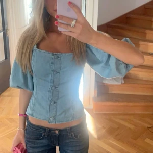 Ljusblå denim blus med puffärmar - Säljer en ljusblå blus i denim med puffiga, korta ärmar och fyrkantig halsringning. Toppen har knappar framtill och rynkade axelband som ger en cool och trendig look. Passformen är figurnära och materialet är mjukt bomullstyg. Perfekt för dig som gillar statement-ärmar! Två små hål som går att sy igen🥹 Mycket eftertraktad därav priset! 😇❣️Lägg rimliga prisförslag ❣️😊