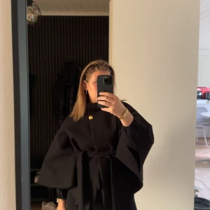 Poncho från missäy  - Säljer en svart kappa med snyggt bälte i midjan och en guldig knapp vid halsen. Jackan har vida ärmar och en stilren, oversized look. Perfekt för dig som gillar minimalistisk och trendig stil.