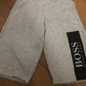 Grå shorts från Hugo Boss XS - Snygga grå shorts från Hugo Boss i storlek XS med stort svart BOSS-tryck längs ena benet. Mjukt material och relaxed passform, perfekta för chill dagar eller träning. Elastisk midja för skön komfort och enkel stil.