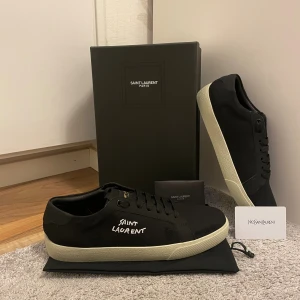 Svarta Saint Laurent sneakers - Svarta sneakers från Saint Laurent med vit broderad logga på sidan och vit platt sula. Skorna har klassisk snörning och är tillverkade i canvas med detaljer i skinn. Perfekta för en clean och stilren look.
