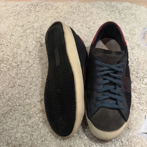 Svarta/blåa Phillipe model, med röd detalj - Snygga svarta sneakers med mörkblå snörning och röda detaljer vid hälen och på sidan. Ovandelen är i mocka och skinn, med en beige platt sula som ger en retrovibe. Perfekta för dig som gillar stilrena men ändå unika skor.