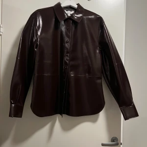 Mörkbrun/vinröd overshirt från H&M Edition - Snygg mörkbrun overshirt från H&M Edition i glansigt syntetmaterial som ger en cool skinnlook. Modellen är loose fit med klassisk krage, långa ärmar och knappar framtill. Perfekt att styla lager på lager för en trendig vibe.