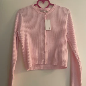 Rosa ribbad kofta från 157 - Supersöt ljusrosa kofta från 157 med ribbad struktur och rund hals. Koftan har långa ärmar och knappar framtill, perfekt för att ge din outfit en mjuk och chill vibe. Croppad passform som sitter snyggt till både jeans och kjol.