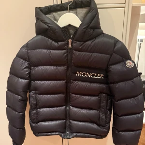 Svart Moncler barn pufferjacka - Svart pufferjacka i storlek 140 från Moncler med broderad logga på bröstet och patch på ärmen. Jackan har en mjuk, fodrad huva i grått, dragkedja framtill, två sidofickor samt en bröstficka. Perfekt för kalla dagar och har en cool, sportig vibe. Köpt på NK i Stockholm. 